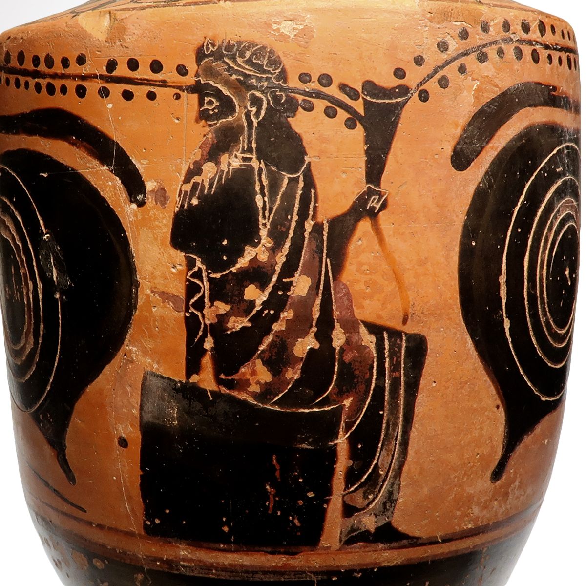 Detail of the scene between the eyes: a bearded figure wearing an ivy wreath sits on a stool, wrapped in a patterned mantle, raising a drinking horn towards a vine with hanging clusters of grapes.